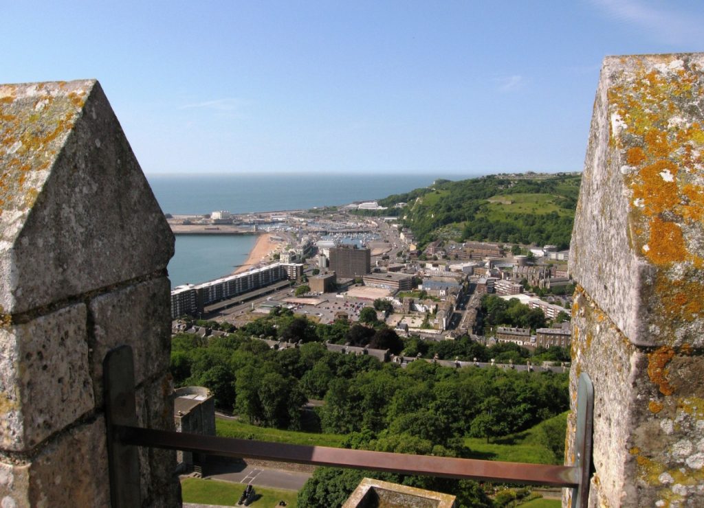 View of Dover from atop the castle, June 2014. The harbour and beach, along with a number of residential buildings and a large car park, are clearly visible. In the background a green hill sits against the sea and a blue sky above.